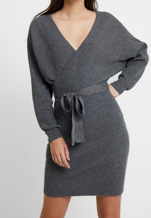 Femme portant une robe en tricot côtelé grise avec manches longues, col en V et ceinture à nouer à la taille, debout avec une main sur la hanche sur fond uni.