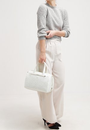 Nainen harmaassa neulepaidassa ja leveälahkeisissa valkoisissa housuissa, kädessään valkoinen Valentino-käsilaukku, jalassaan mustat teräväkärkiset korot, seisoo yksiväristä taustaa vasten.