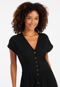 Robe noire à boutons avec un décolleté en V profond, des manches courtes et un tissu texturé. Elle présente une taille cintrée qui s'évasent au niveau de la jupe.