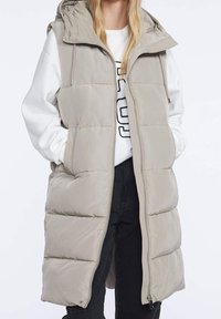 Beige gesteppte Weste mit Kapuze, Reißverschluss vorne und gefütterten Taschen. Mit gesteppten Einsätzen für Struktur und einem entspannten Schnitt. Wird über einem weißen Sweatshirt getragen.