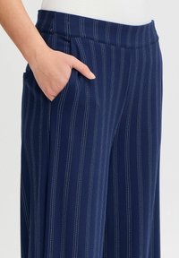 Pantalons rayés marine en tissu lisse, avec des rayures pinstripes blanches verticales, une taille élégante et des poches latérales.