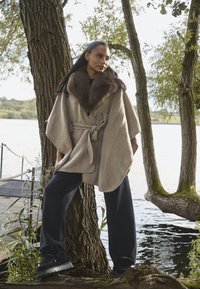 Poncho en mélange de laine beige avec un large col en fourrure marron, ceinturé à la taille, associé à un pantalon noir à jambes larges et des chaussures sombres.