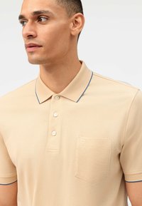 Beige polo shirt van een gestructureerde stof, contrasterend met een kraag met blauwe afwerking en een borstzak met een geborduurd logo. Vijfknops sluiting.