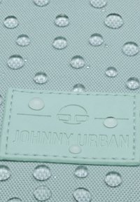 Kapky vody na texturované tkanině obklopující šitý obdélníkový štítek s vyraženým logem a textem "JOHNNY URBAN".