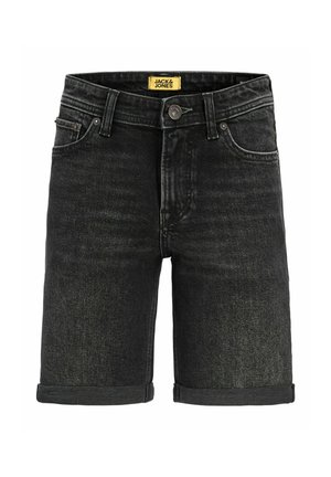 Gutt står, iført lyseblå genser, svarte oppbrettede denimshorts, hvite sokker og beige sko, mot ensfarget hvit bakgrunn.