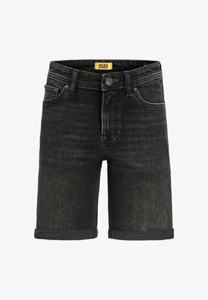 Fiú áll, világoskék pulóvert, fekete felhajtott farmer rövidnadrágot, fehér zoknit és bézs cipőt visel, egyszínű fehér háttér előtt.