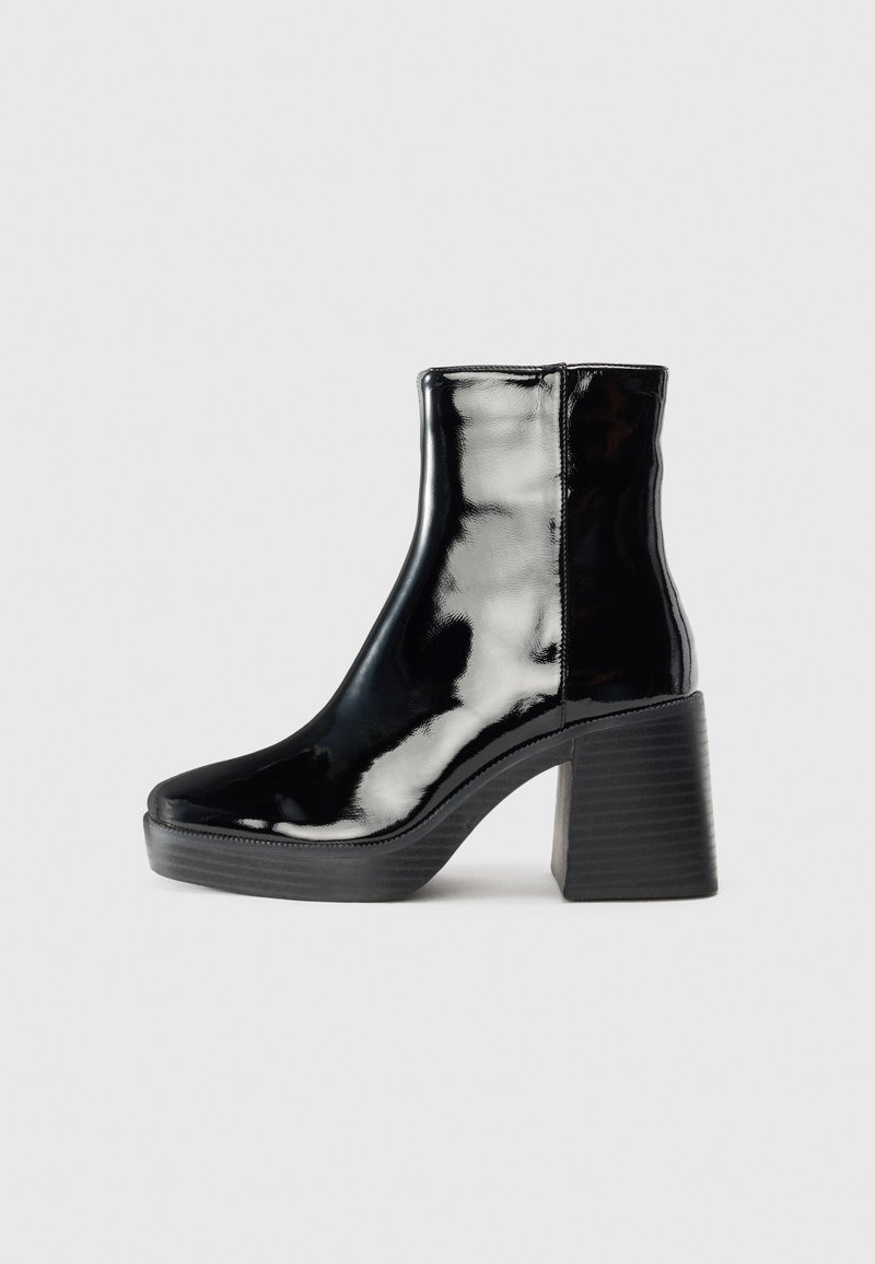 Bottine noire brillante à cheville avec talon bloc épais et bout légèrement arrondi sur fond gris clair uni.