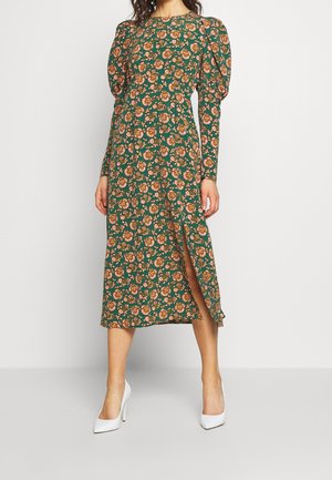 Žena v zelených midi šatech s růžovými a oranžovými květinovými vzory, nafouklými dlouhými rukávy, předním rozparkem a bílými vysokými podpatky.