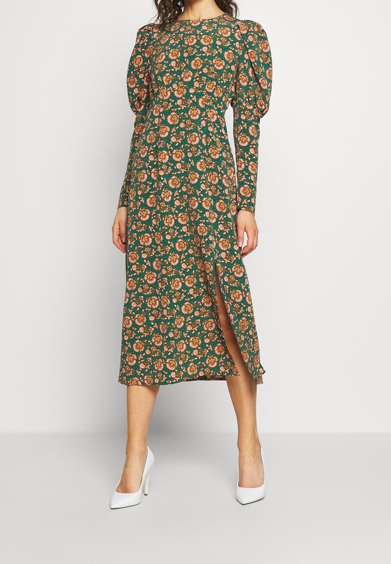 Femme portant une robe midi verte à motifs floraux roses et orange, avec des manches longues bouffantes, une fente à l'avant, et des chaussures blanches à talons hauts.