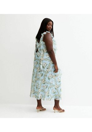 Woman in a blue floral midi dress with ruffled straps and open back, wearing beige low-heeled sandals, standing against a plain white background.