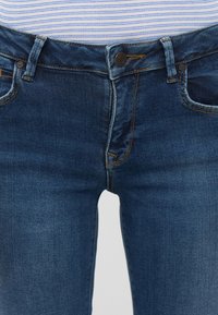 Σκούρα μπλε τζιν παντελόνια με στενή γραμμή, διαθέτοντας πέντε τσέπες, πορτοκαλί ραφές και μεταλλικό κούμπωμα στη μέση.