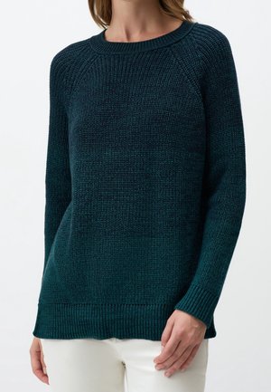 Femme portant un pull en tricot à manches longues bleu sarcelle foncé, associé à un pantalon blanc, debout devant un fond clair uni.