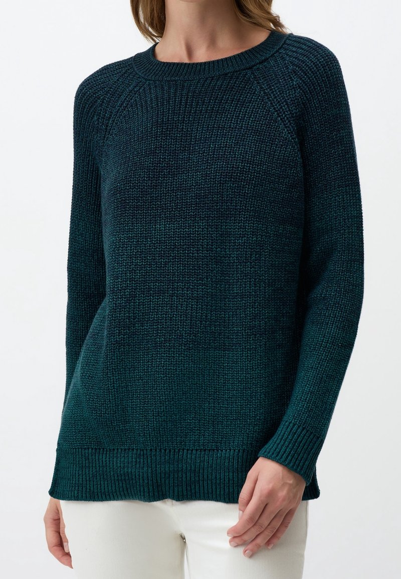 Femme portant un pull en tricot à manches longues bleu sarcelle foncé, associé à un pantalon blanc, debout devant un fond clair uni.