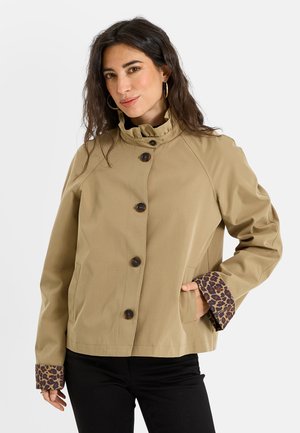 Mujer con cabello oscuro que lleva una chaqueta beige abotonada con puños de estampado de leopardo y pantalones negros, posando con una mano en el bolsillo frente a un fondo blanco.