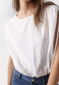 Top bianco a maniche corte con tessuto testurizzato caratterizzato da piccoli punti ricamati, vestibilità comoda e ampia scollatura, abbinato a jeans in denim blu.