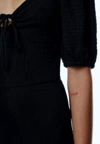 Schwarz texturiertes Oberteil mit Kordelzug-Ausschnitt und Puffärmeln. Am Arm ist ein rotes Tattoo mit dem Jahr "1964" zu sehen.