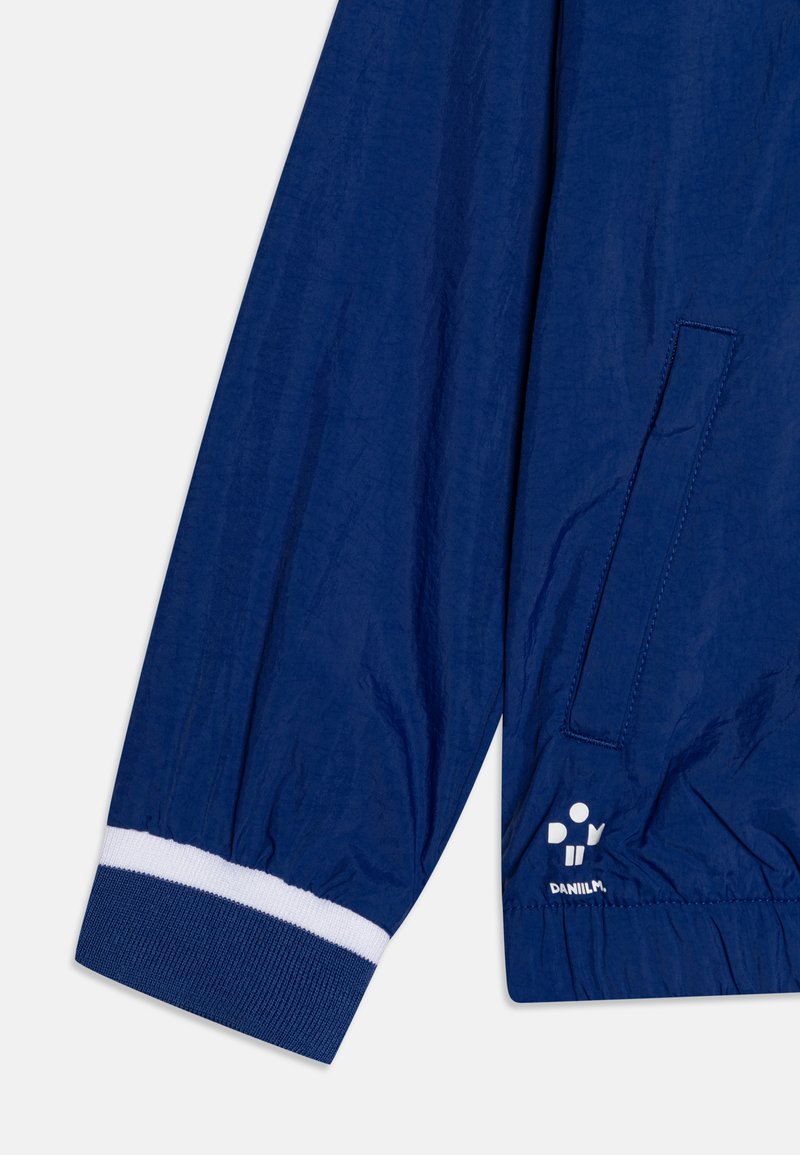 Veste bleu marine avec des poignets et une taille élastiques. Présente une poche et des accents blancs. Le matériau est léger avec une finition légèrement texturée.