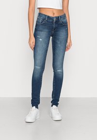 Jeans de ganga azul skinny com áreas desbotadas, design de cinco bolsos e detalhes de costura subtis. Combinados com ténis brancos.