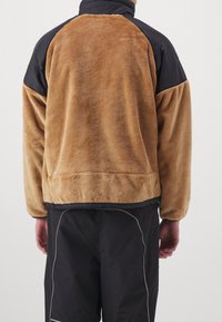 Giacca in pile con corpo beige, pannelli neri sulle spalle e colletto alto. Materiale morbido e texturizzato con orlo e polsini elasticizzati.