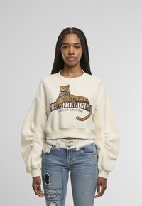 Sudadera corta de color crema con un gráfico de leopardo y el texto "True Religion Brand Jeans". Combinada con jeans azules desgastados y accesorios de plata.