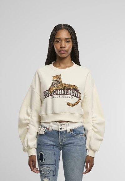 Sweatshirt court crème avec un graphique de léopard et le texte "True Religion Brand Jeans". Associé à un jean bleu déchiré et des accessoires en argent.