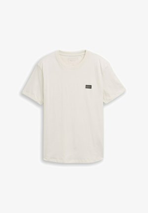 Jednostavna majica kratkih rukava krem boje s uskim okruglim izrezom i malim pravokutnim crnim logotipom na lijevoj strani prsa.