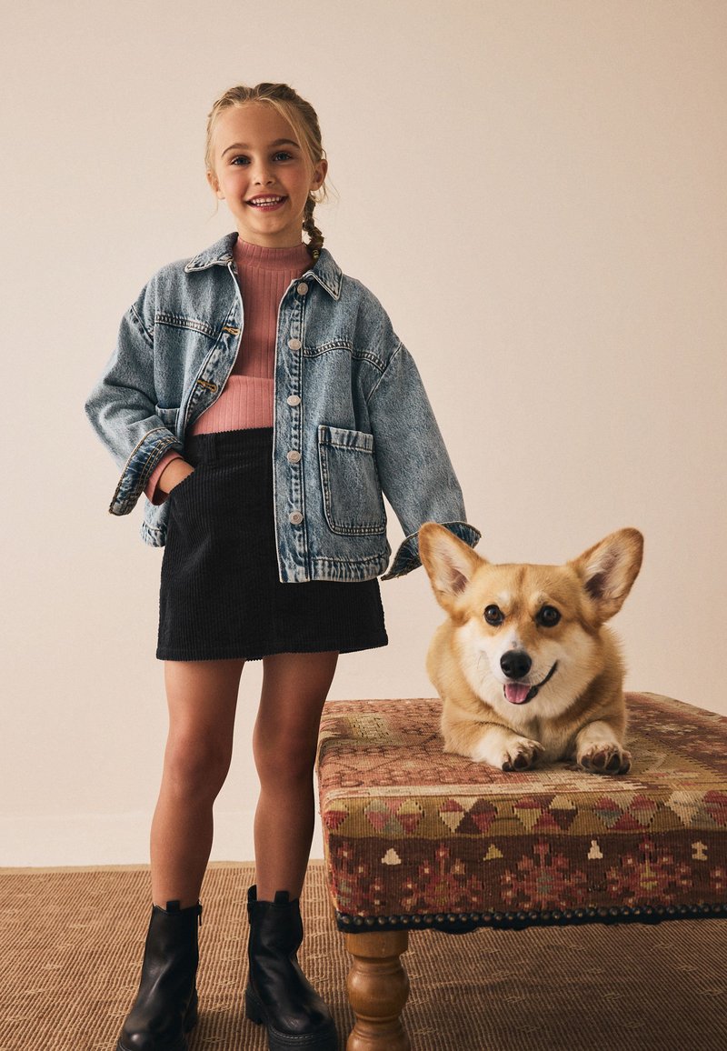 Giacca di jeans, top rosa a coste, gonna nera di velluto a coste e stivali neri. Corgi accanto alla ragazza su una panchina decorata con accenti colorati.