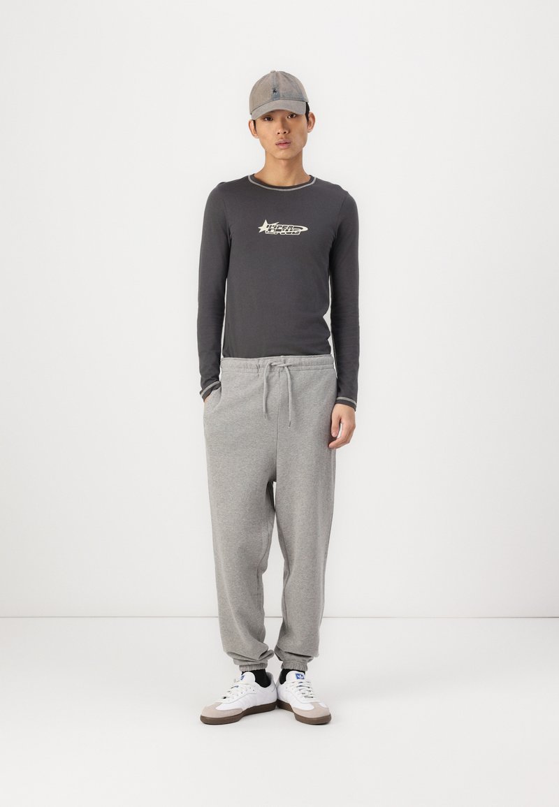 Zwarte langemouwen shirt met een grafisch logo, gecombineerd met lichtgrijze trainingsbroek en witte sneakers met grijze accenten, gedragen met een grijze pet.