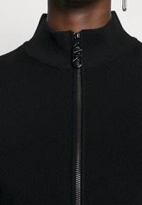 Top nero a coste con colletto alto e chiusura con zip anteriore, caratterizzato da un tirante della zip in nero lucido per un tocco elegante.