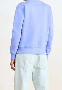 Person som bär en ljuslila sweatshirt och ljus tvättade denimjeans, sedd bakifrån mot en enkel ljus bakgrund.