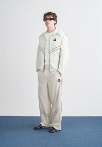 Ανοιχτόχρωμο μπουφάν με φερμουάρ και μαύρη διακόσμηση, συνδυασμένο με μπεζ παντελόνι με μαύρες ρίγες. Το μοντέλο φοράει μαύρα γυαλιά ηλίου και παπούτσια.