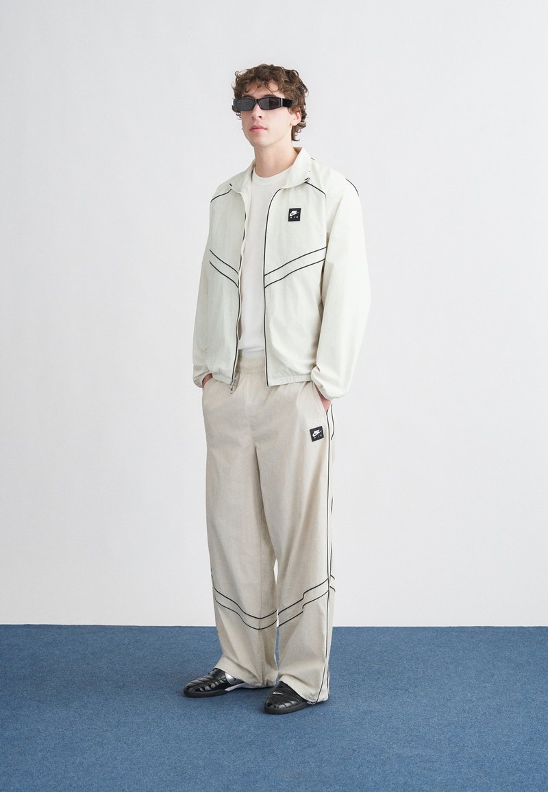 Chaqueta de cremallera clara con ribetes negros, combinada con pantalones beige con rayas negras. El modelo lleva gafas de sol negras y zapatos negros.