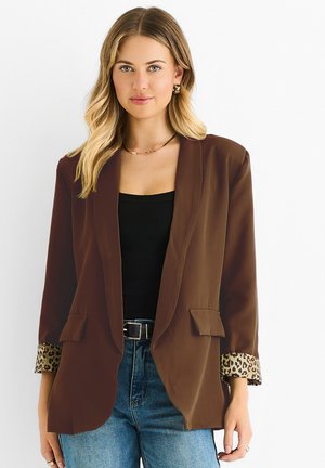 Young woman with blonde hair wearing a brown blazer with leopard print cuffs, black top, blue jeans, and gold jewelry against a white background.