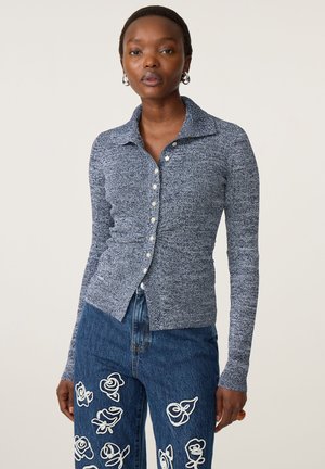 Vrouw draagt een aansluitende grijze, met knopen gesloten trui en blauwe jeans met witte bloemrijke borduur, staand tegen een effen achtergrond.