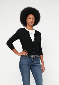 Moteris su afro šukuosena, vilkinti juodą meuotą kardiganą, baltą marškinėlį, mėlynas džinsus ir juodą diržą, stovi viena ranka ant klubo.