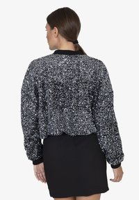 Giacca bomber nera con paillettes, caratterizzata da una superficie texture, polsini e orlo a coste. Indossata su una gonna nera aderente, che mette in mostra un design corto.