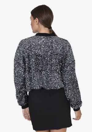 Giacca bomber nera con paillettes, caratterizzata da una superficie texture, polsini e orlo a coste. Indossata su una gonna nera aderente, che mette in mostra un design corto.