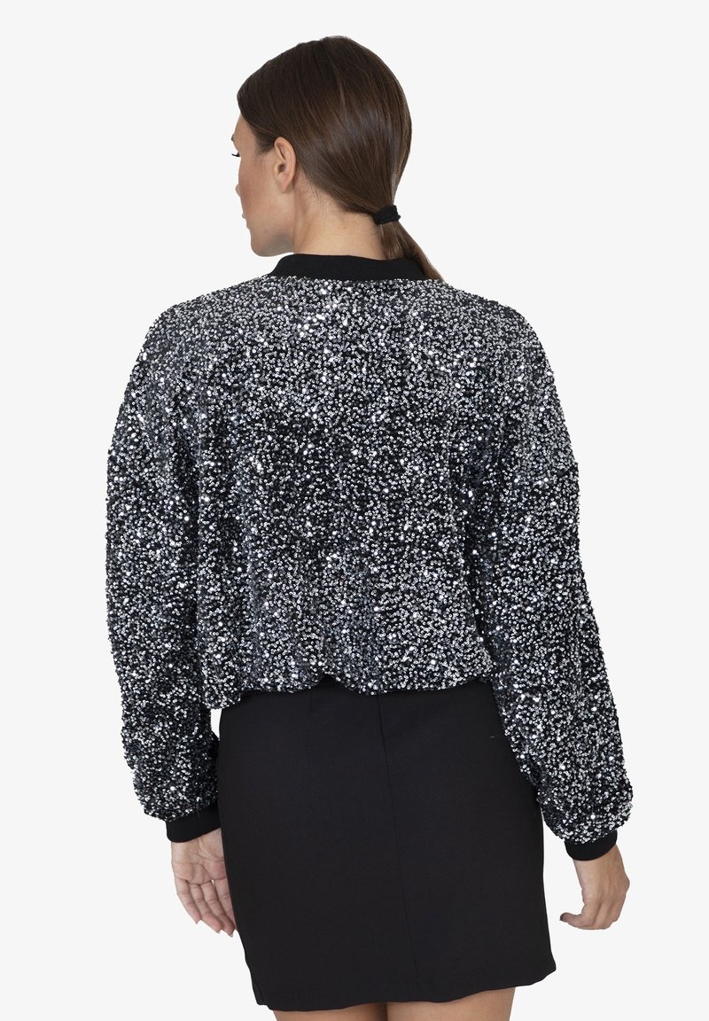 Giacca bomber nera con paillettes, caratterizzata da una superficie texture, polsini e orlo a coste. Indossata su una gonna nera aderente, che mette in mostra un design corto.