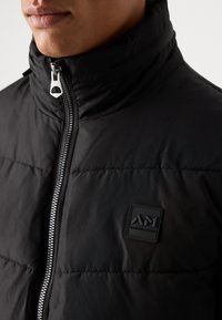 Chaqueta acolchada negra con cuello alto, con una cremallera plateada y un parche de logo texturizado en el frente. Tejido suave con diseño acolchado.