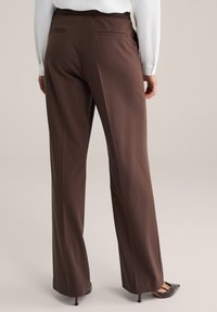 Vrouw draagt hoge bruine wijd uitlopende broek met achterzakken met bies, gecombineerd met een wit shirt met lange mouwen en donkere hakken.