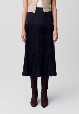 Jupe midi plissée noire avec une taille lisse, des panneaux texturés contrastés et un ourlet fluide ; associée à des bottes hautes en daim marron.