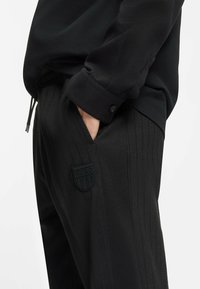 Schwarze Hose mit Nadelstreifenmuster und einem gummierten HUGO-Logo-Patch am linken Oberschenkel. Hergestellt aus strukturiertem Stoff, mit Seitentaschen.