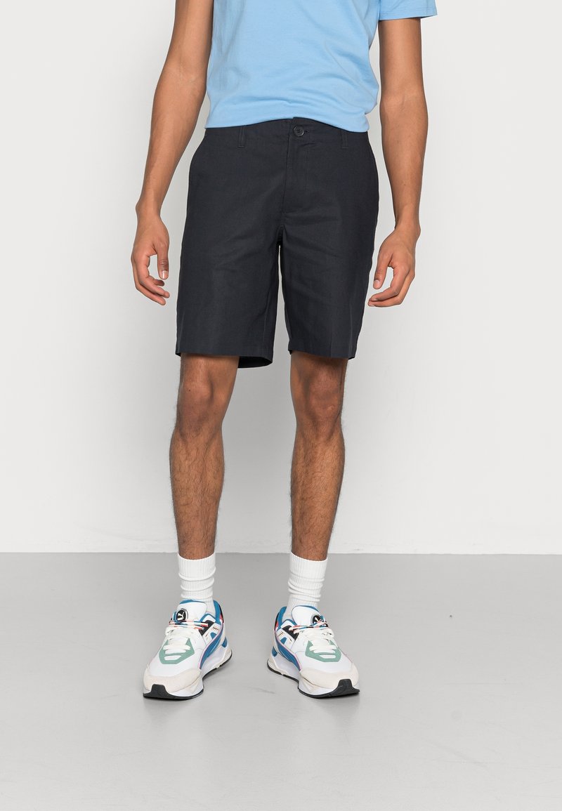Shorts bleu marine en tissu mélangé coton, dotés d'une fermeture à bouton, de poches latérales et d'une coupe droite, associés à un T-shirt bleu clair et des baskets multicolores.