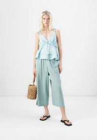 Svetlomodrý top s peplum strihom a volánovým lemom, skombinovaný s voľnými, svetlozelenými culottes. Modelka drží tkanú slamennú tašku a má na sebe čierne šľapky.