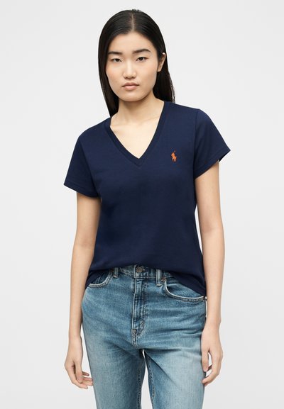 Γυναίκα που φοράει μπλε σκουρό μπλουζάκι πόλο με V-λαιμόκοψη και πορτοκαλί λογότυπο, σε συνδυασμό με ανοιχτό μπλε τζιν, στέκεται μπροστά σε λευκό απλό φόντο.