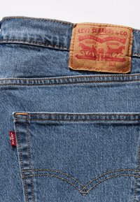 Jeans de denim azul con un parche de cuero marrón que muestra "Levi Strauss & Co." en texto rojo, con costuras naranjas y una pequeña etiqueta de logo roja.