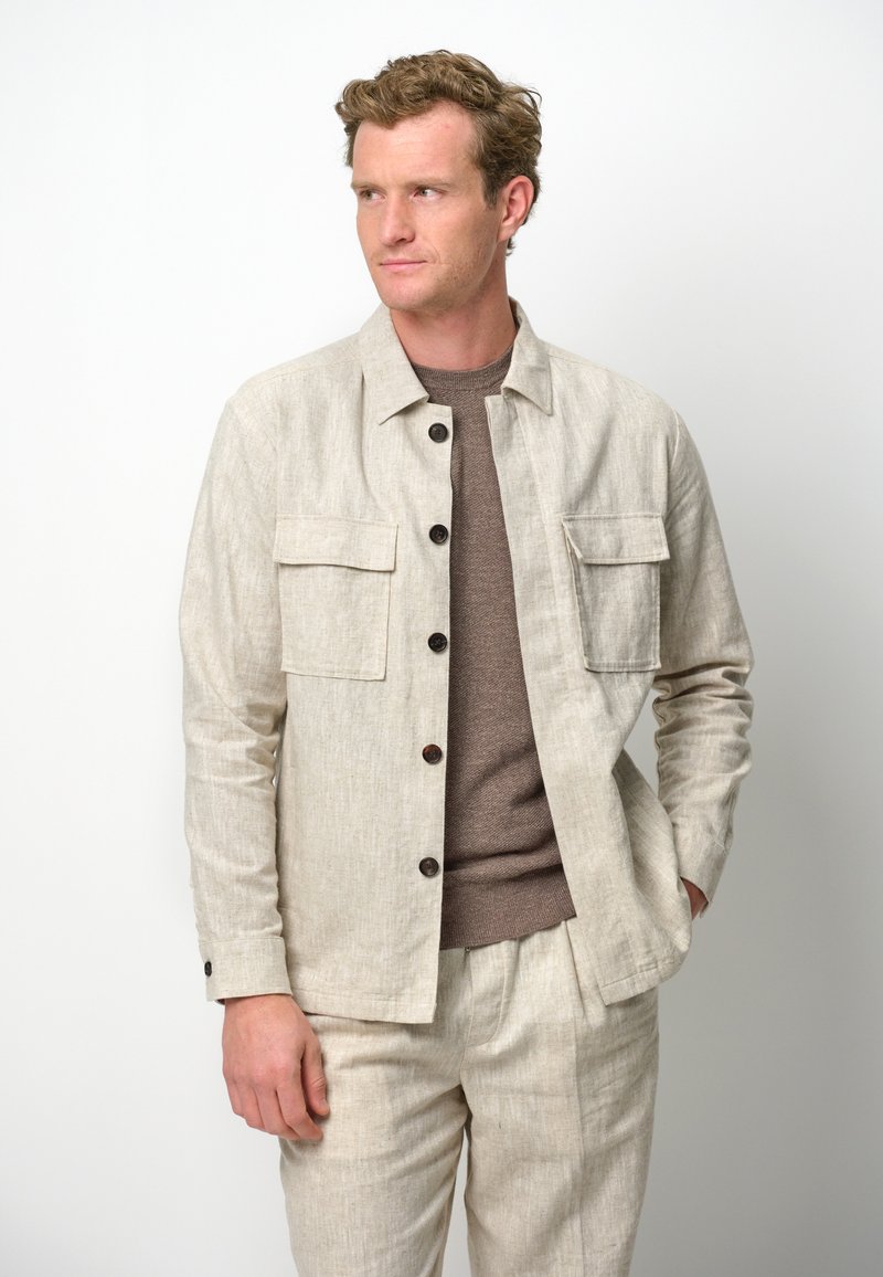 Giacca di lino beige chiaro con due tasche anteriori e bottoni marroni, indossata sopra un maglione marrone texture, con una vestibilità sartoriale.