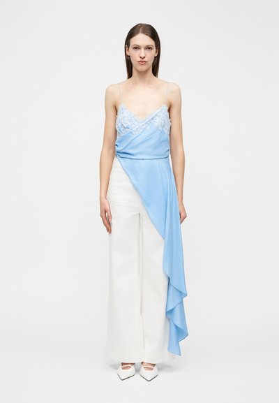 Femme portant un haut asymétrique bleu clair avec des bordures en dentelle et un tissu drapé, associé à un pantalon blanc à jambes larges et des chaussures blanches à bouts pointus.