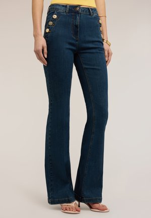 Femme portant un jean évasé bleu foncé avec des boutons dorés décoratifs sur les poches avant et des sandales blanches à talons.