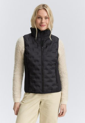 Vrouw met blond haar, gekleed in een zwart doorgestikt vest over een beige textuurtrui en lichtgekleurde broek, tegen een effen achtergrond.